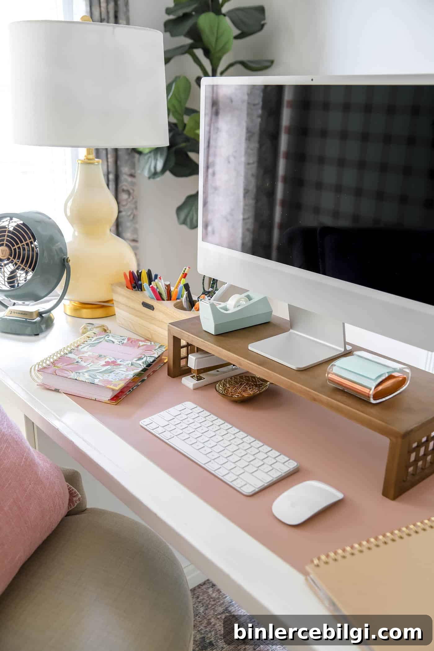 organized desk space with mac computer and desk riser, fan and lamp.