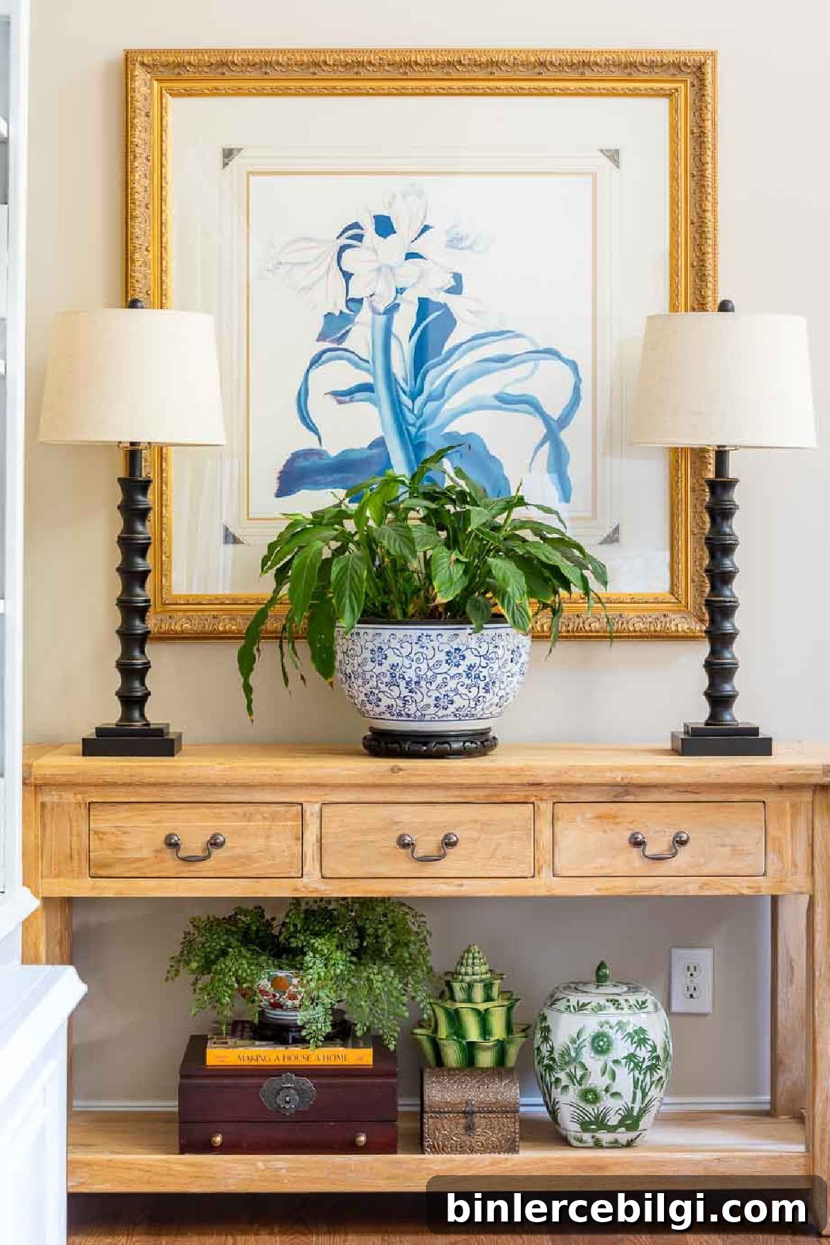 wood console table with lamps, wall art behind it and a large potted plant on the middle of the table.