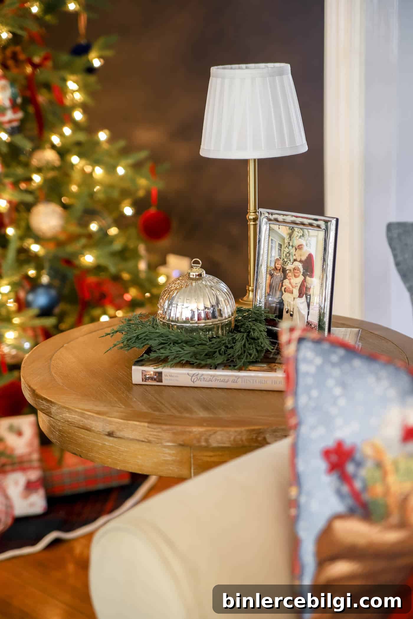 side table with Santa and children in a frame, small cordless lamp and ornament candle with a christmas coffee table book and greenery.
